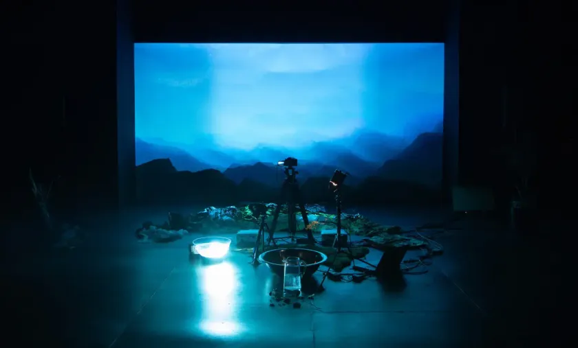 Performance documentation from askîwan: a darkened stage lit in blue, a projected mountain landscape behind a camera on a tripod, with bowls of water and scattered material on the floor.