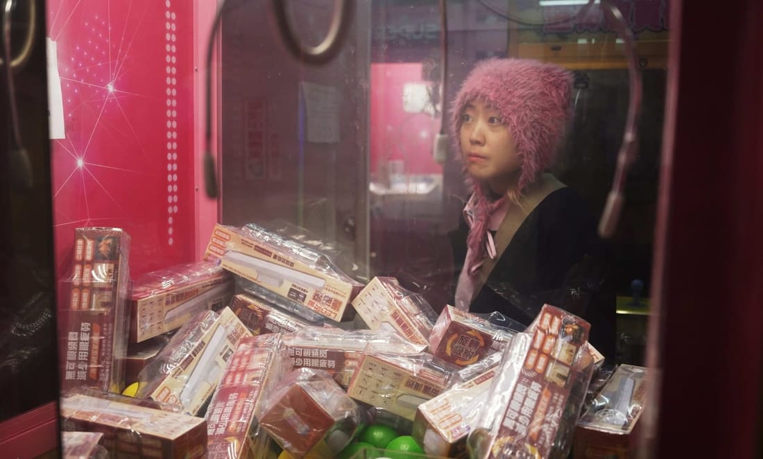 Performance documentation from [guāng yīn] at MAI, 2025: a performer in a pink fur hat seen through the glass of a claw machine packed with boxed prizes.